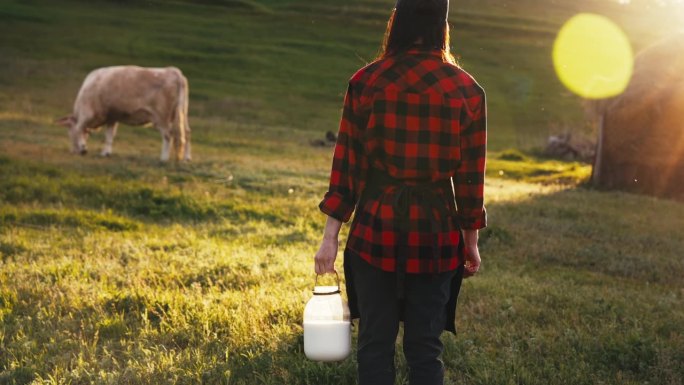 妇女站在牧场牧场的牧场上，手里拿着一个牛奶罐给牛挤奶。为农牧生产天然、健康的产品。夏天的阳光照在大自