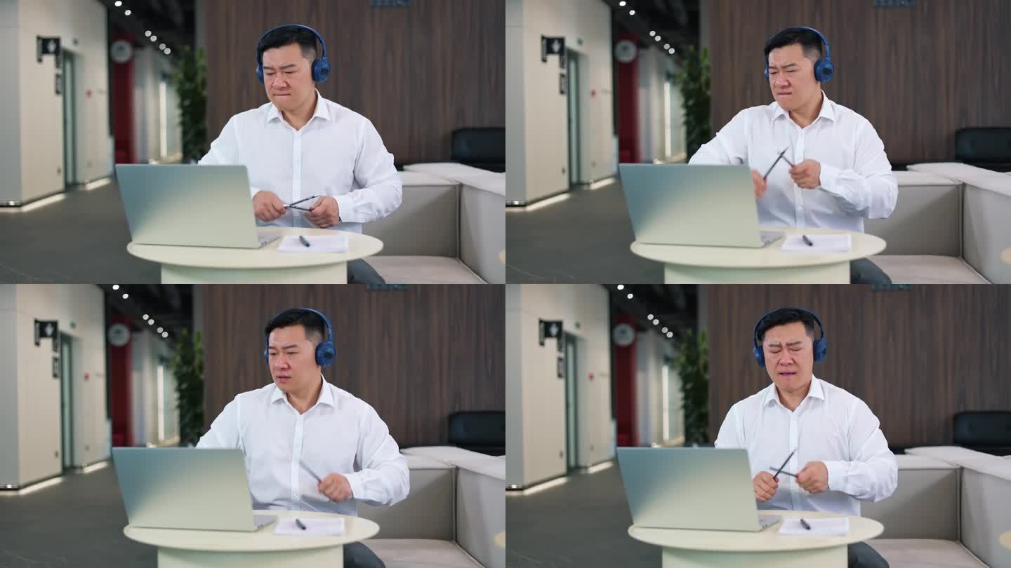 成功的男性企业家戴着无线耳机听着歌，看着笔记本电脑屏幕，用铅笔模仿打鼓。积极的亚洲人在现代办公室休息