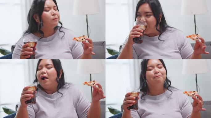 大码亚洲女人享受披萨和软饮料，而放松在家里的沙发上。