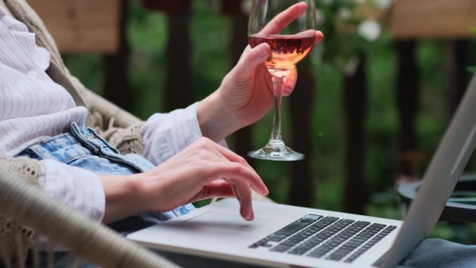 一个女人手拿着一杯玫瑰酒，在舒适的家庭露台上使用笔记本电脑。女性的手指在触摸板上滑动。家庭办公，远程