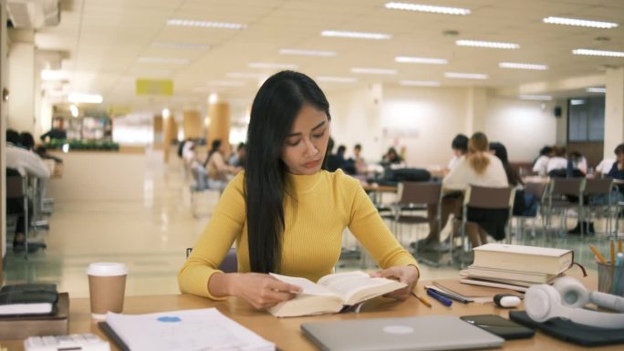 亚洲学生女孩坐在桌边一边看书一边做作业，用笔记本电脑在国外通过网友联系打视频电话，在图书馆里开心地笑