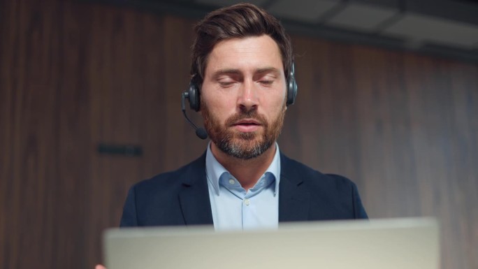 穿着西装的自信的白人男子在工作场所使用无线耳机和笔记本电脑进行视频会议。留胡子的英俊男企业家在现代办