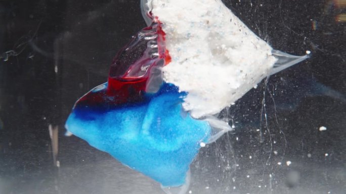 为洗碗机准备的可溶解包装在水下溶解的特写镜头。红色凝胶和蓝白色粉末在水中慢慢释放和分散。