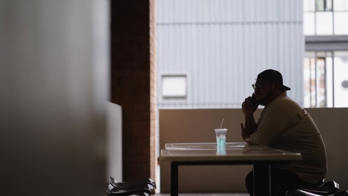 一名体重超标的男学生在短暂的休息时间，坐在大学阳台的长椅上，桌上放着一杯冰苏打水，一边用手机和朋友聊