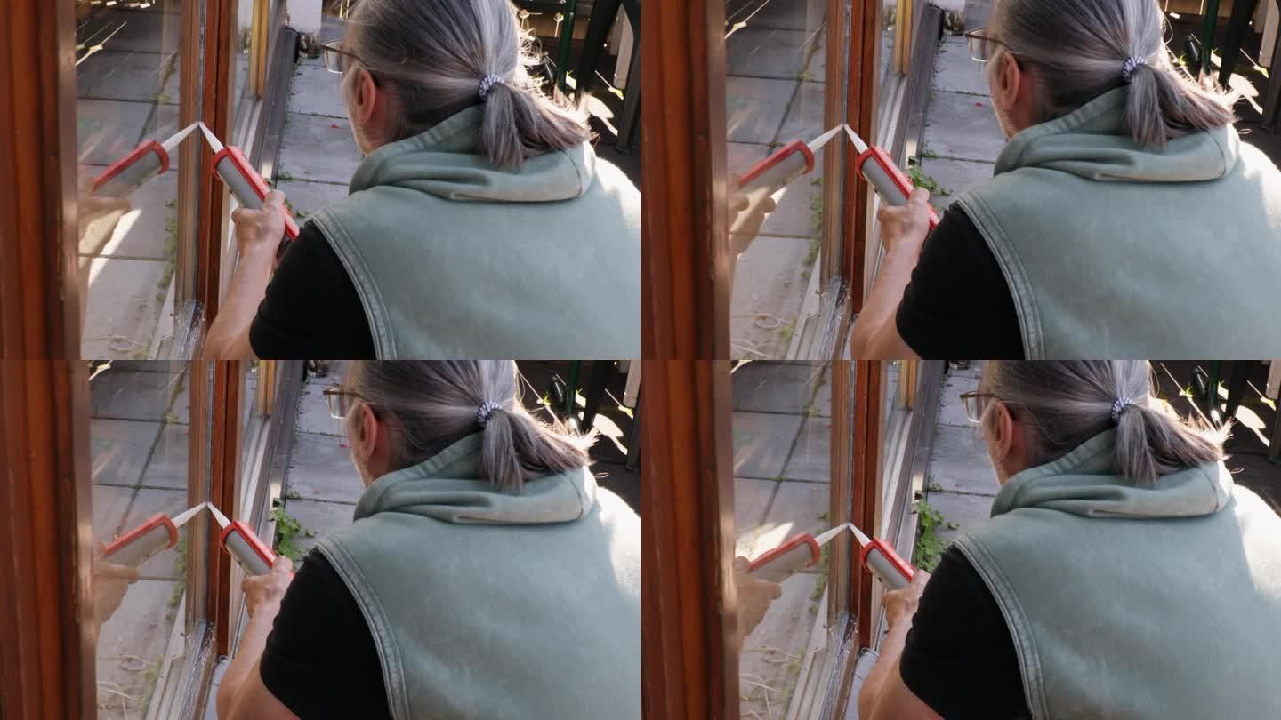 一个头发花白、扎着马尾的成年男子用硅胶填塞阳台门上的裂缝