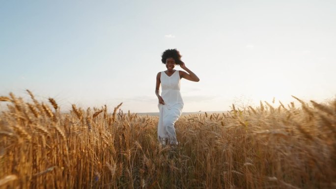 旅行的概念。优雅的黑人妇女，留着一头白色的长发，穿着白色的连衣裙，穿过麦田，迎着风微笑着，在夏日的夕