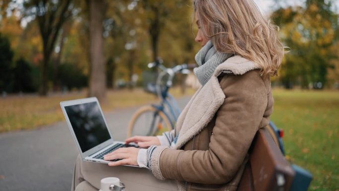 在秋天的公园里，穿着时髦保暖衣服的金发女郎坐在长椅上使用笔记本电脑