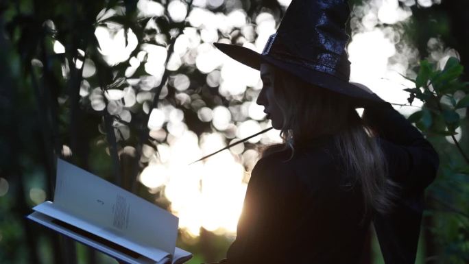 幻想女女巫手里拿着一本魔法书念咒语。美丽的女巫在常绿的森林里吟诵咒语。