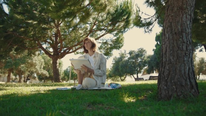一个成年女子坐在绿色的公园里，一边在速写本上画画，一边聊天