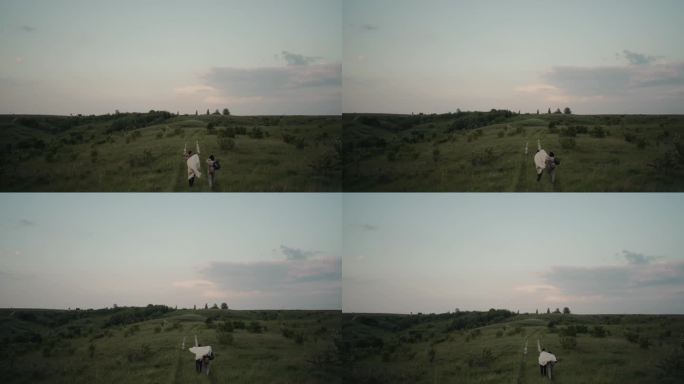 夕阳下，一对年轻的夫妇披着毯子，拥抱着走在夏日的田野里。浪漫的慢动作镜头。