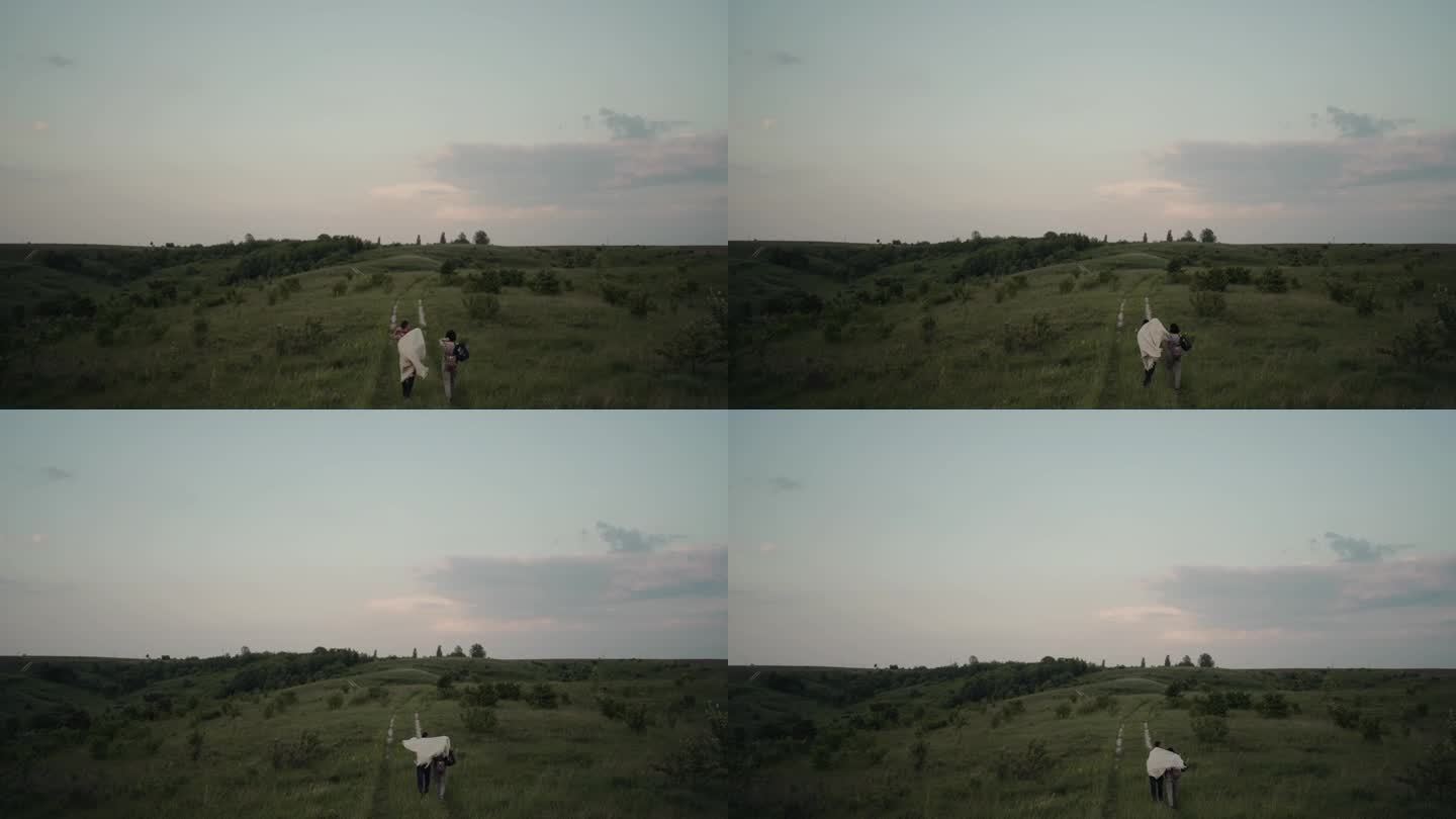 夕阳下，一对年轻的夫妇披着毯子，拥抱着走在夏日的田野里。浪漫的慢动作镜头。