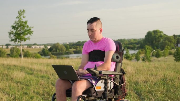 坐在轮椅上的男子在草地上用现代笔记本电脑工作