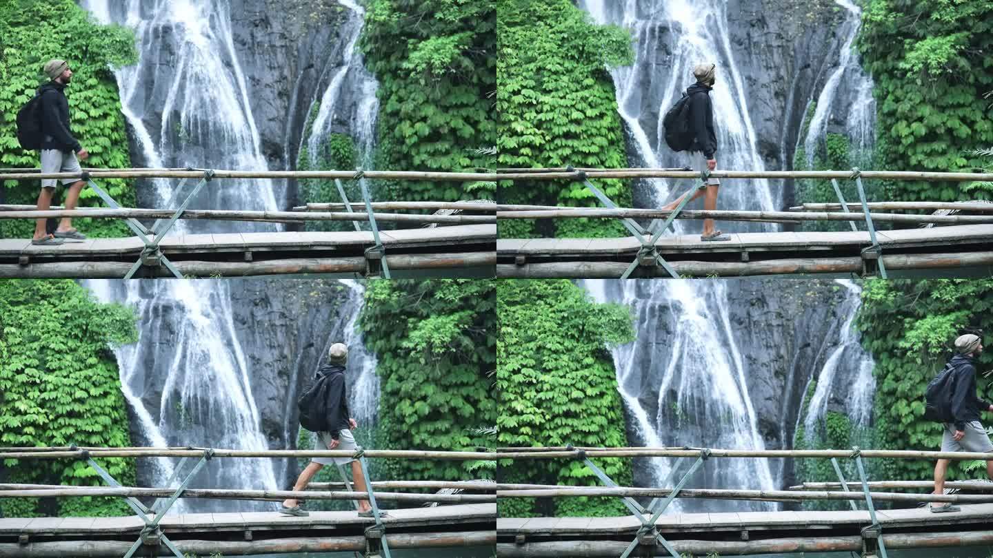成年男子在绿色自然中前往森林瀑布。从桥上走到丛林瀑布目的地的年轻人的生活方式。郁郁葱葱岛天堂之旅的概