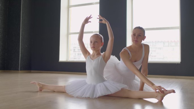 小芭蕾舞女演员坐在地板上，老师在她身后