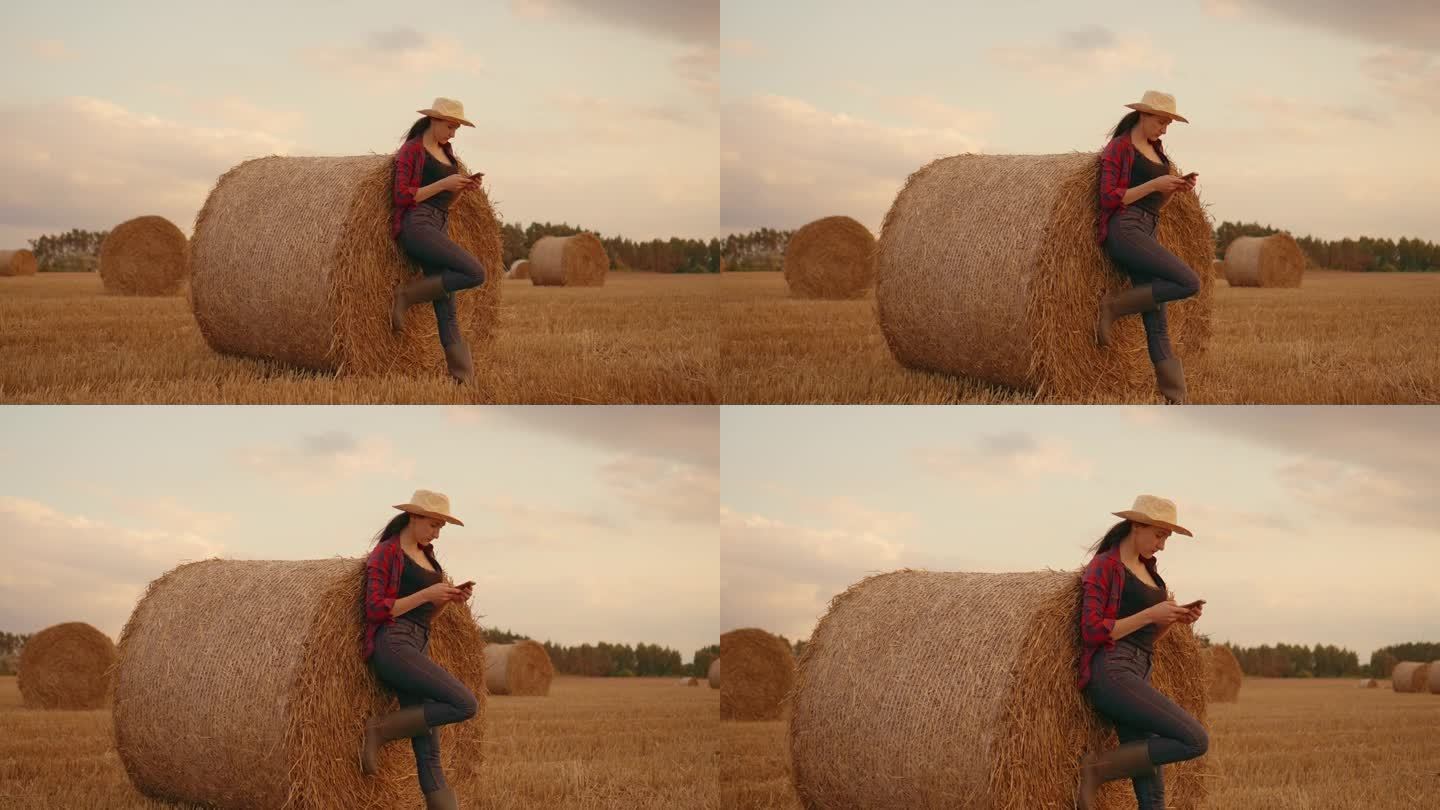 牛仔风格的女人穿着橡胶靴，格子红衬衫，戴着草帽，站在田野里的圆草堆旁，用着手机
