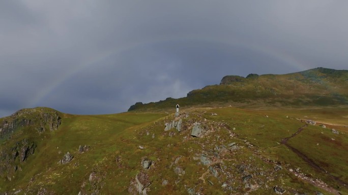 无人机飞向站在悬崖顶上的年轻男性旅行者，观察峡湾上方美丽的彩虹