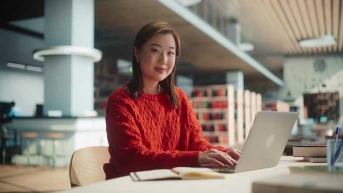漂亮的日本女学生在图书馆的笔记本电脑上写大学学位论文。年轻的亚洲女人看着镜头，微笑着。穿着休闲红色套