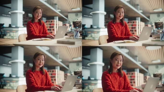 漂亮的日本女学生在图书馆的笔记本电脑上写大学学位论文。年轻的亚洲女人看着镜头，微笑着。穿着休闲红色套