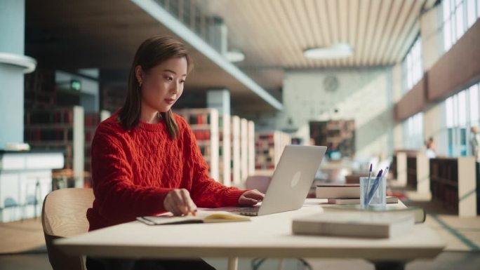 在现代公共图书馆阅览室，一位身着红色大衣的亚洲女性为课堂作业创作专题计划，在笔记本上写作，并使用笔记