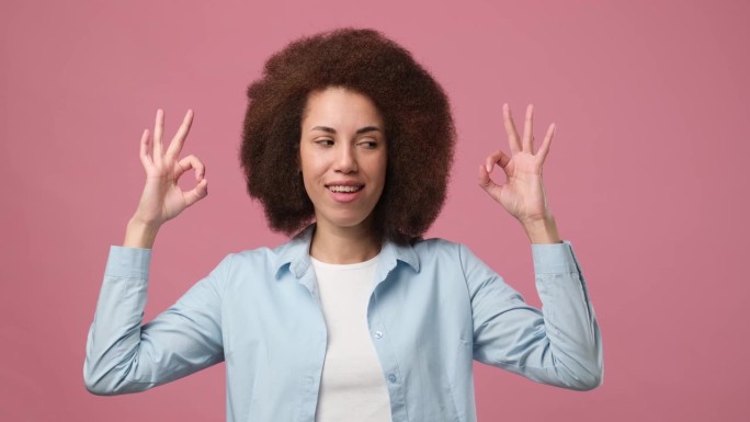 开朗的年轻非洲裔美国女孩，留着非洲发型，点头示意，用手指站在粉红色的工作室背景上