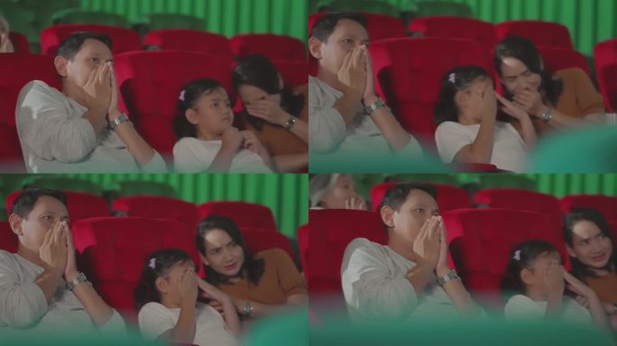 在现代电影院看恐怖电影的亚洲家庭，在黑暗的大厅里被吓得用手捂住眼睛。不同种族的人在电影院看电影。集体