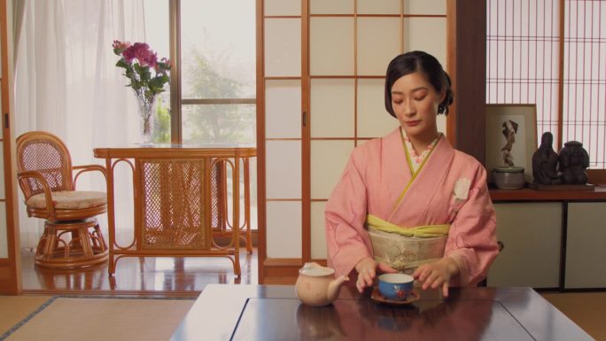 年轻的日本女性享受独自在家喝茶的时光。漂亮的亚洲女人穿着经典的粉红色和服，坐在传统日本家庭的矮桌子后