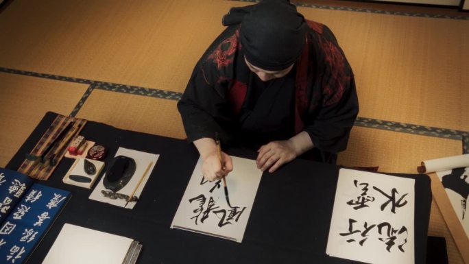 一位日本艺术家为客户委托书法作品的高角度镜头。成人书法家坐在传统的工作室里，用墨水在纸上书写汉字符号