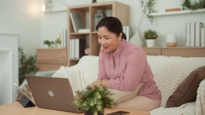 一位亚洲成年女性正坐在沙发上通过笔记本电脑观看网上娱乐节目，而她的女儿正端着一个茶杯给她。