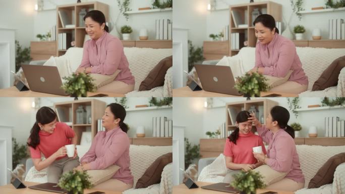 一位亚洲成年女性正坐在沙发上通过笔记本电脑观看网上娱乐节目，而她的女儿正端着一个茶杯给她。