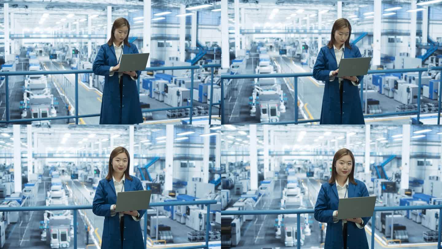 一个年轻的亚洲技术人员使用笔记本电脑和看着相机的肖像。在一家现代电子厂工作的穿着蓝色外套的工业女工