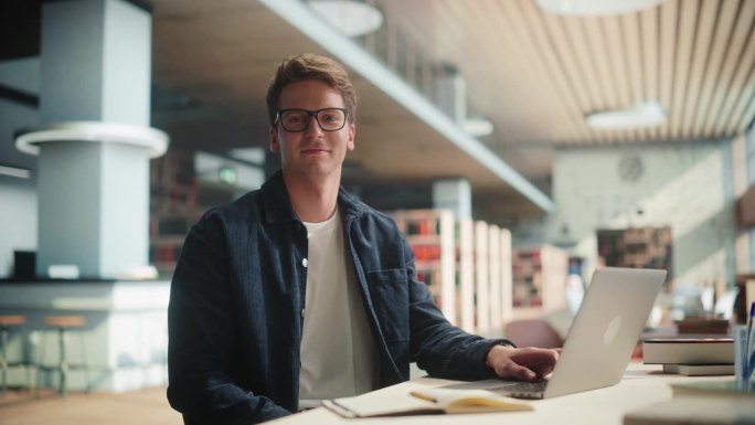 一个帅气的男学生在现代图书馆的笔记本电脑上做他的大学作业。年轻的白人男子微笑着看着镜头。他戴着眼镜，