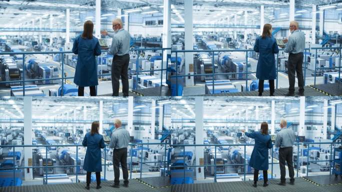 男女工程师背对着相机站着，使用笔记本电脑，在一个工厂里交谈，工厂里有为不同行业生产现代电子元件的设备
