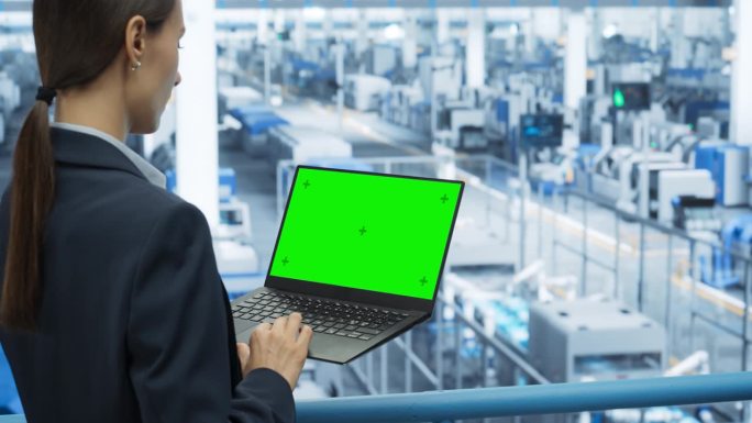 年轻的女工程师在电子厂使用绿屏模拟显示的笔记本电脑。越过肩膀的镜头。自动化制造机器和人工智能机器人被