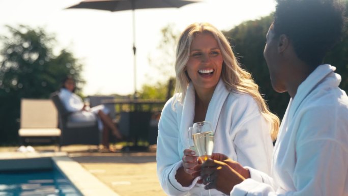 两个成熟的女性朋友穿着长袍，在室外泳池边喝香槟