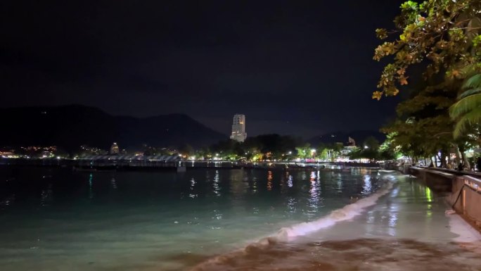 夜晚的泰国普吉岛芭东海滩，美丽的白色沙滩和蓝绿色的海水上灯火通明。远处的闪电和烟花