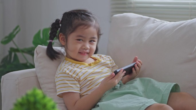 亚洲小女孩坐在沙发上看手机屏幕。宝宝笑逗的时候用手机。看屏幕时间太长。可爱的女孩一边看电视一边看视频
