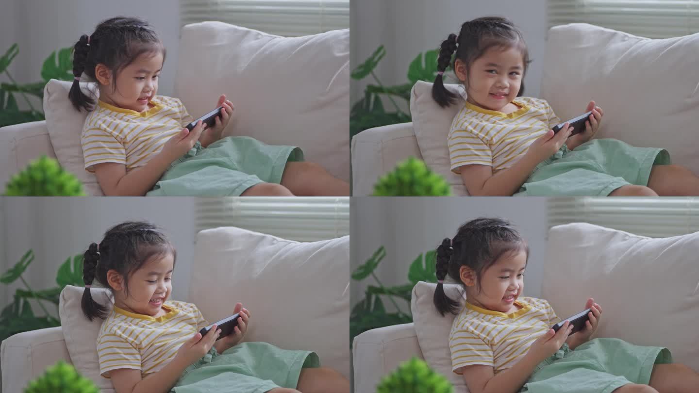 亚洲小女孩坐在沙发上看手机屏幕。宝宝笑逗的时候用手机。看屏幕时间太长。可爱的女孩一边看电视一边看视频