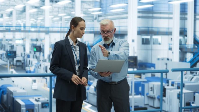 两个不同的工程师站在一个电子工厂的平台上，使用笔记本电脑。机器在生产电子元件，专家在监控在线软件的进