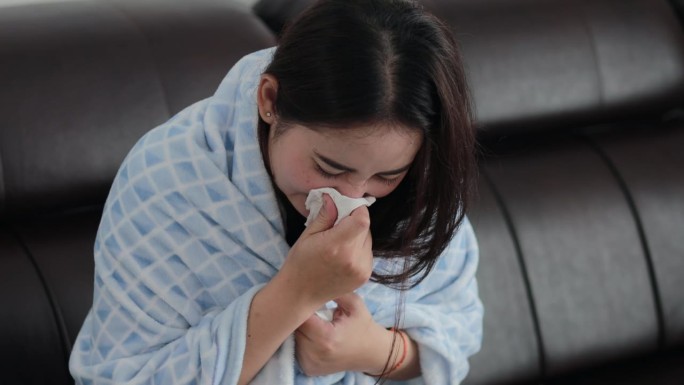 不健康的亚洲妇女裹着毯子吹感冒打喷嚏流鼻涕过敏流感咳嗽感冒打喷嚏。