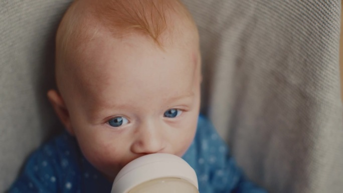 可爱的小男孩的高角度肖像享受奶瓶里的牛奶，同时放松在家里的保镖。蓝色的眼睛