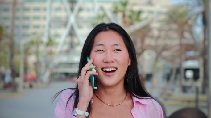 慢动作镜头，一个亚洲女人一边走一边用智能手机友好地交谈。中国漂亮的女学生微笑着用手机聊天。女孩用手机