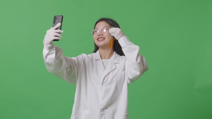 亚洲女科学家拿着一试管橙色液体，站在实验室的绿色屏幕背景上，用智能手机打视频电话