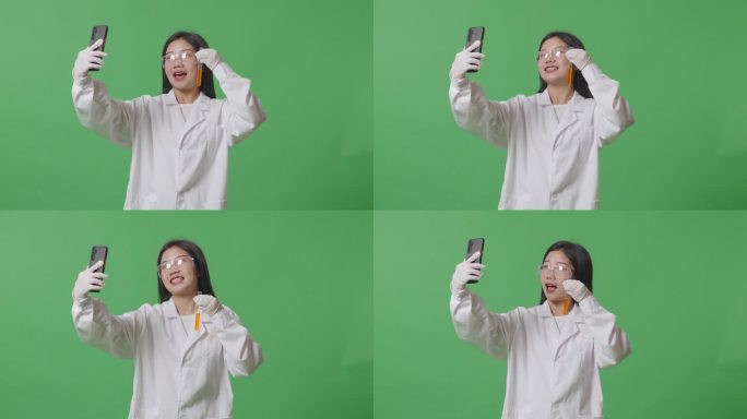 亚洲女科学家拿着一试管橙色液体，站在实验室的绿色屏幕背景上，用智能手机打视频电话