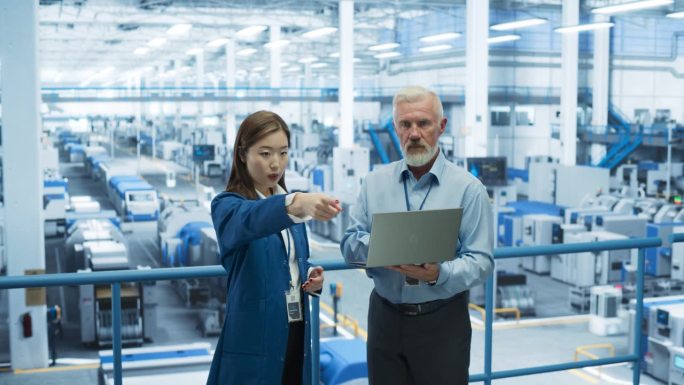 一个中年工程师和快乐的亚洲科学家的肖像，在一个工厂里使用笔记本电脑和谈话，工厂里有为不同行业生产现代