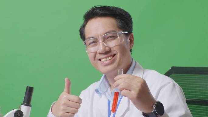 在绿屏背景的实验室里，亚洲男性科学家一边看着试管，一边微笑着竖起大拇指，一边用显微镜在桌子上工作