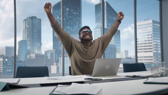 黑人商人坐在摩天大楼办公室的办公桌前，用笔记本电脑工作，情绪激动地庆祝电子商务交易的成功。非裔美国企