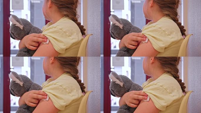 注射疫苗后，白人女孩抱着泰迪熊坐在诊所候诊室里，用纱布垫压着针头止血