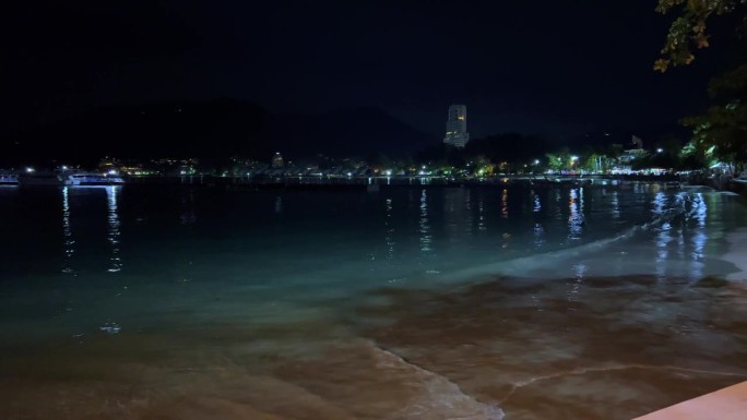 夜晚的泰国普吉岛芭东海滩，美丽的白色沙滩和蓝绿色的海水上灯火通明。远处的闪电和烟花