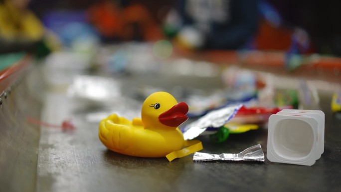 特写黄色橡皮鸭在传送带上与垃圾和模糊的工人分类垃圾在慢动作背景。工厂的玩具和垃圾在室内回收。