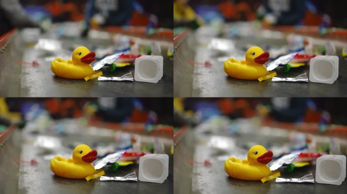 特写黄色橡皮鸭在传送带上与垃圾和模糊的工人分类垃圾在慢动作背景。工厂的玩具和垃圾在室内回收。